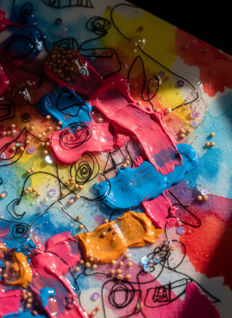 A detailed macro photograph of an interdisciplinary art surface by Studioviralsneha: thick acrylic brushstrokes layered over translucent watercolor washes, with tiny beads and iridescent sequins embedded in clear gel medium. Fine black ink lines dance across the surface, weaving between raised textures and glossy patches. The color palette pulses with neon pinks, electric blues, and warm oranges. A single directional studio light from the right creates dramatic highlights on glossy areas and soft shadows in crevices, emphasizing dimensionality. Photographic realism with extreme close-up framing, capturing just a small section of the artwork so it feels abstract and immersive. The atmosphere is bold, experimental, and tactile, drawing attention to the handcrafted detail.